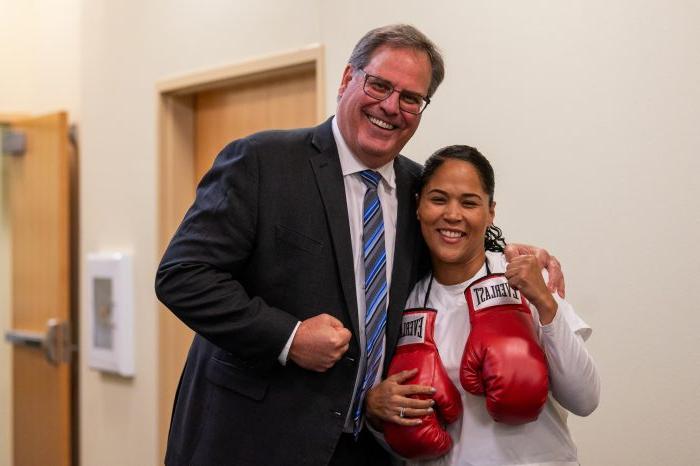 A woman with boxing gloves hanging from her shoulders poses with a man as both jokingly display fists for the camera.